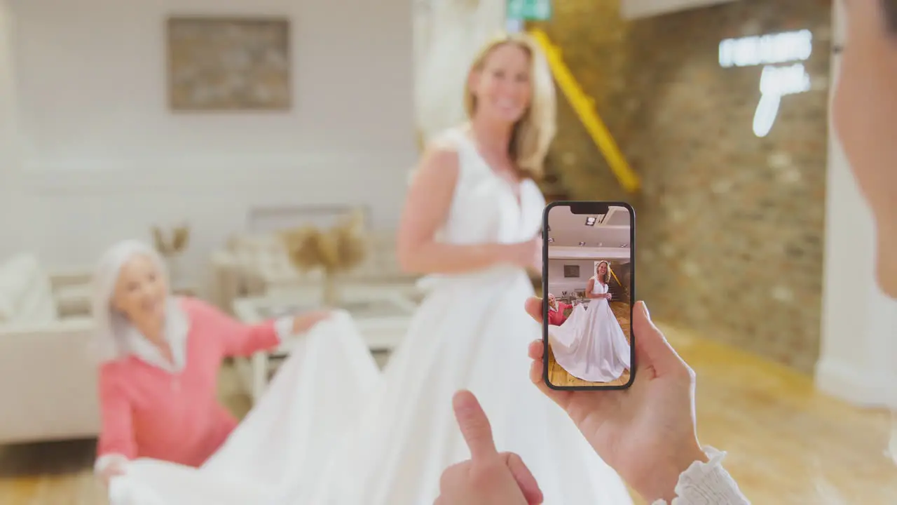 Grandmother With Adult Daughter And Granddaughter Trying On Wedding Dress Taking Picture On Phone