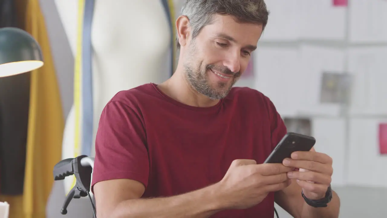 Mature Male Fashion Designer In Wheelchair In Studio Checking Text Messages On Mobile Phone