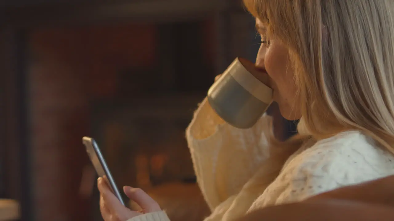 Close Up Of Woman At Home In Lounge With Cosy Fire And Hot Drink Using Mobile Phone