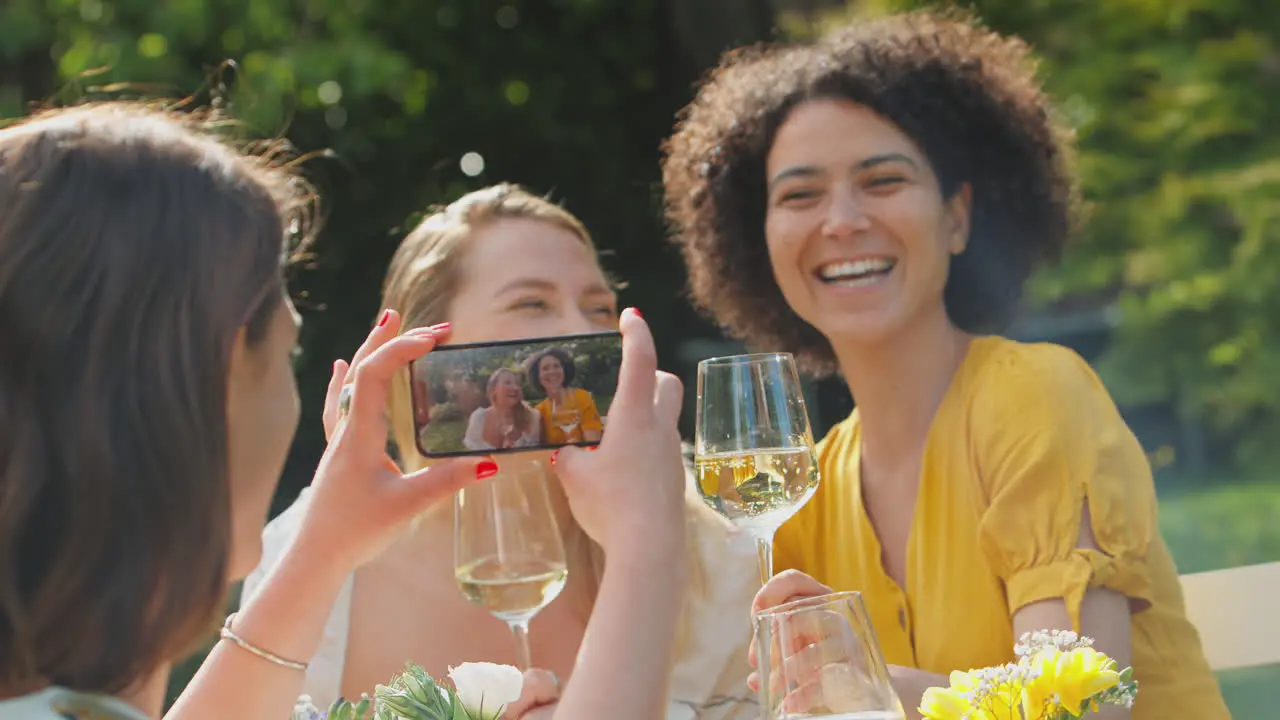 Woman Taking Video Of Friends Sitting Outdoors In Summer Garden Drinking Wine And Making A Toast