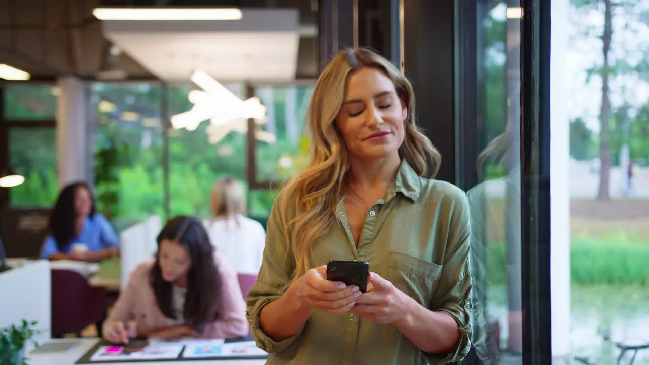 Mature Businesswoman Standing By Window In Busy Office Texting Or Browsing On Mobile Phone