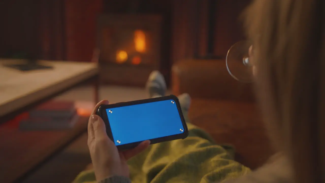 Woman At Home Sitting On Sofa With Fire Streaming To Blue Screen Mobile Phone Holding Glass Of Wine