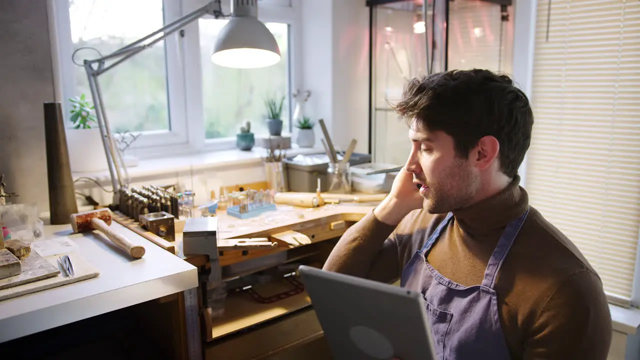 Male Jeweller Making Phone Call Looking At Design On Digital Tablet In Studio