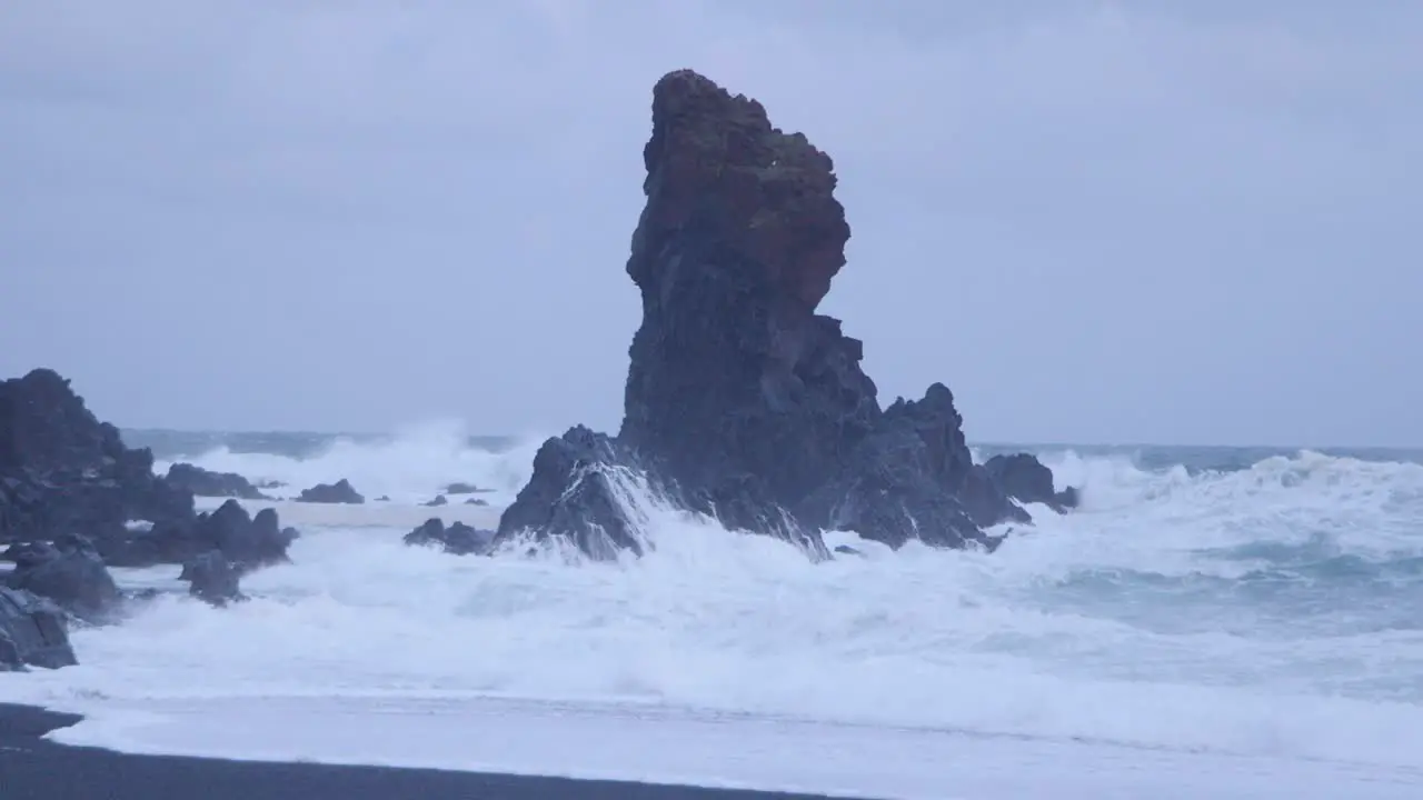 Dramatic waves crashing against unique rock formations on a black sand beach in Iceland under overcast skies