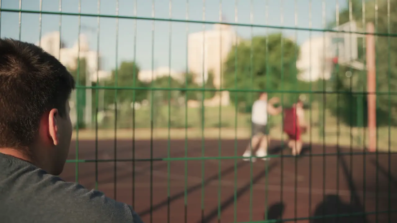 Back View Of A Young Man Sitting And Watching Basketball Training Session In An Outdoor Basketball Court In A Sunny Day 1