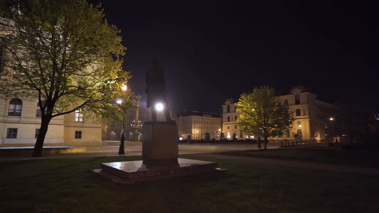 A statue of a person on an empty plaza in Prague Czechia at night during a Covid-19 lockdown trees and the magnificent concert hall of Rudolfinum in the background light fog over the square pan