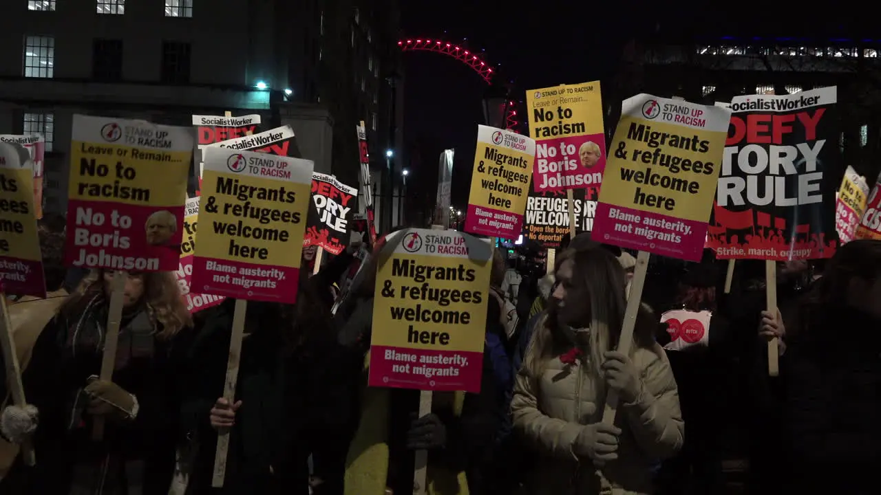 Anti-racist protestors stand on Whitehall at night holding Stand Up To Racism and Socialist Worker placards in front of a lit up London Eye during a protest against Prime minister Boris Johnson