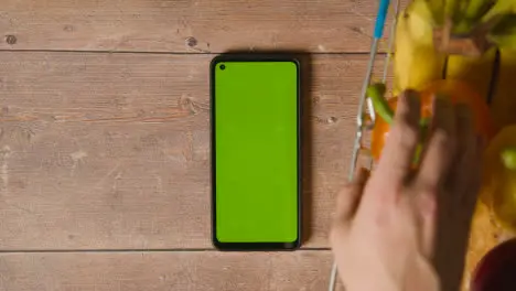 Overhead Studio Shot Of Person Picking From Basic Food Items In Supermarket Wire Shopping Basket Next To Green Screen Mobile Phone 