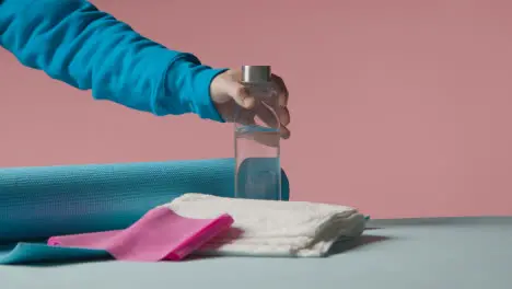 Fitness Studio Shot Of Person Picking Up Water Bottle With Exercise Mat And Towel Against Pink Background