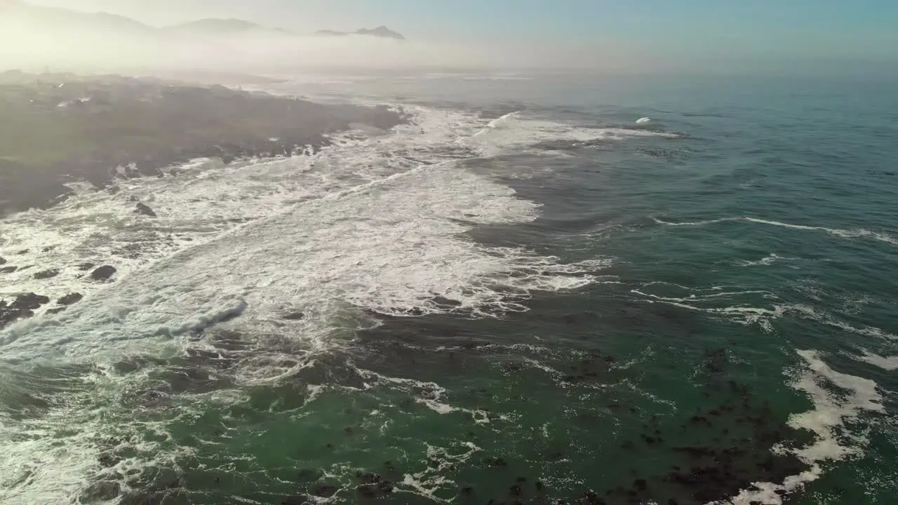 High flying aerial view of ocean and land as waves roll in while panning up from waters to display shoreline with a bit of mist