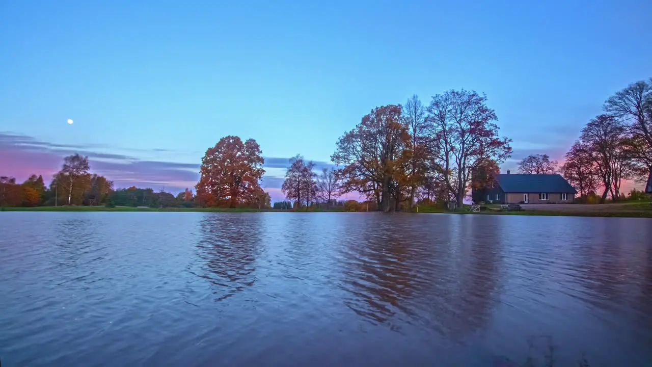 The moon descends as the sunrise illuminates the fall-color trees by a lake and cabin time lapse