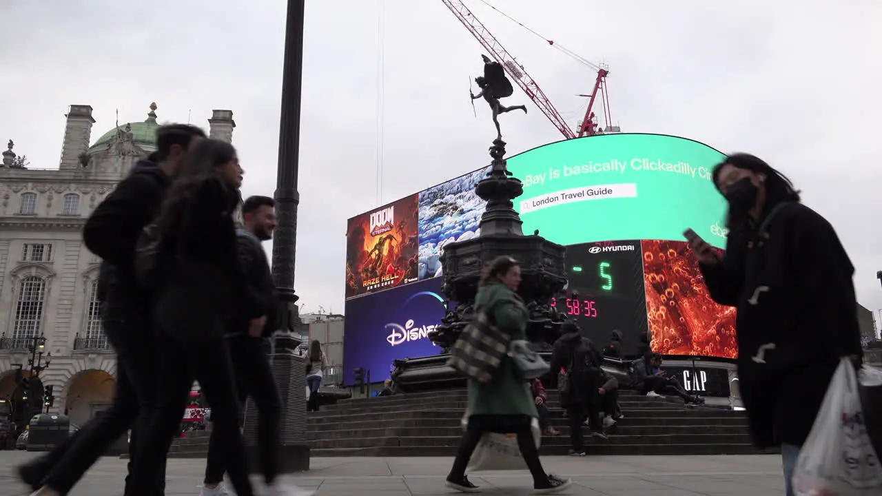 A woman wearing a surgical face mask walks past the Eros Statue and the neon advertisement billboard sign at Piccadilly Circus