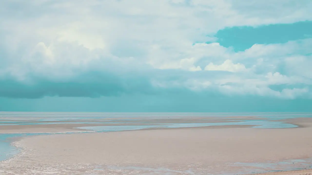 Timelapse of large storm clouds brewing on the horizon of a beach at lowtide where large veins of water stretch along the pale sand whilst the warm sun still beams