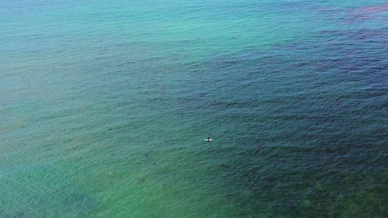 A lone surfer sits on his surfboard waiting for a wave like a spec in the vast ocean surface aerial view