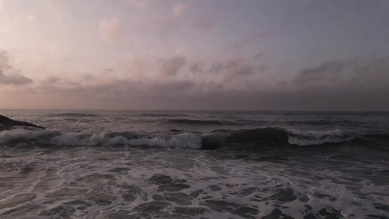 Low angle aerial pull back across dramatic sunset rocky splashing coastal beach skyline