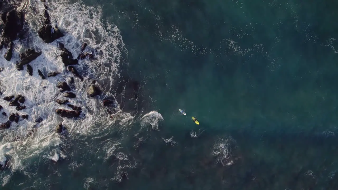 Two surfers wating for waves near rocky coastline top down zoom in drone view