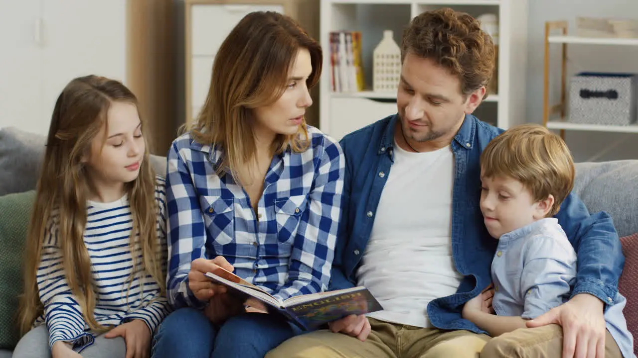 Cheerful Family Of Parents And Little Son And Daughter Sitting All Together On The Couch And Flipping The Pages Of The English Book