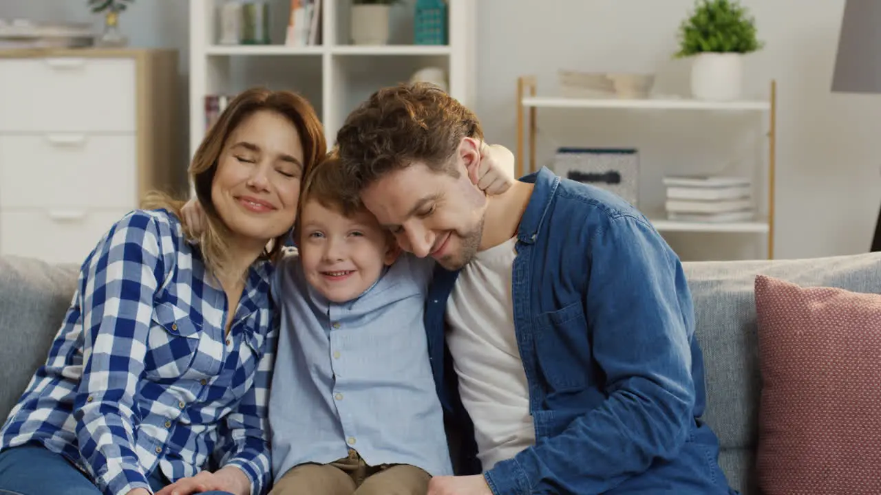 Portrait Shot Of The Young Attractive Parents Sitting With Their Little Cute Son On The Sofa In The Living Room Hugging And Smiling To The Camera