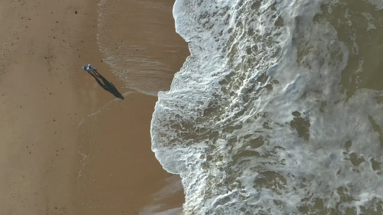 Aerial shot of parent and child social distancing while walking on a beach with waves crashing