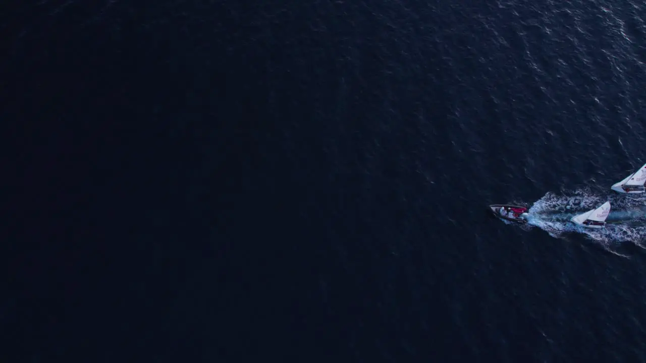 Motorboat towing four white sail boats returning to shore at nightfall top down aerial