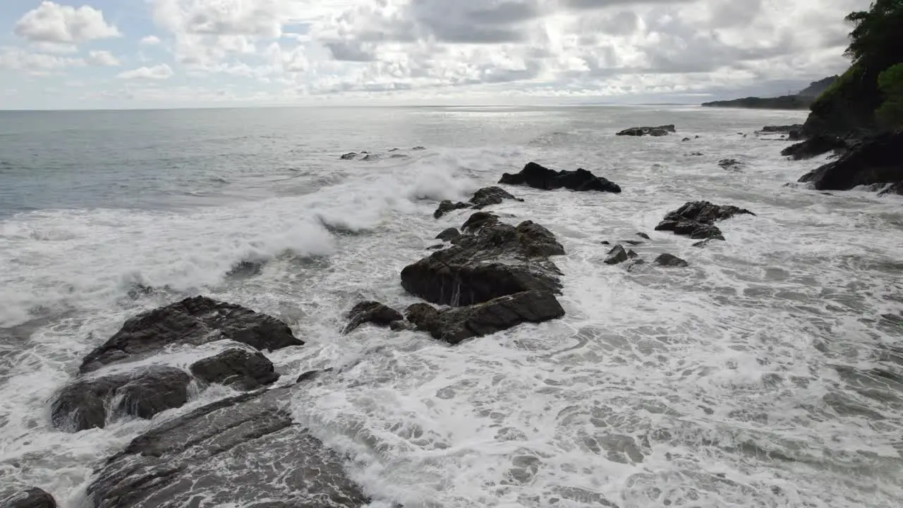 Aerial dolly in flying over foamy sea waves hitting the rocky shore on a cloudy day in Dominicalito Beach Costa Rica