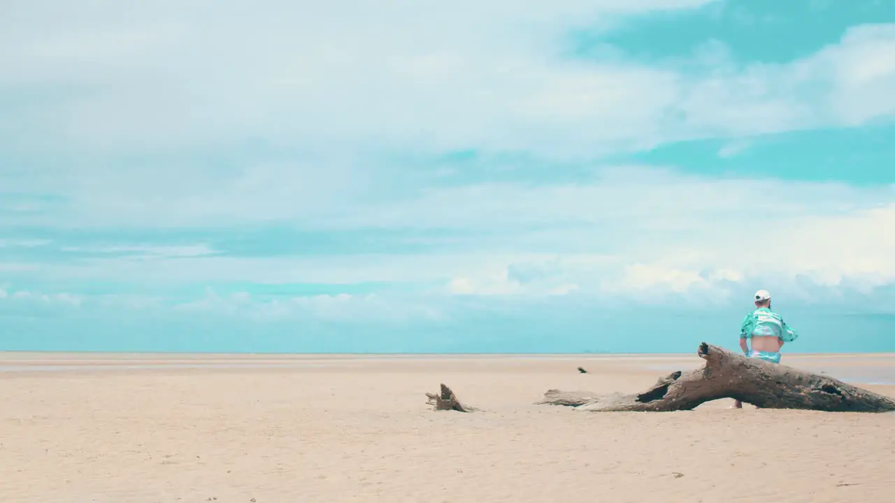 A young man with a beard sits on a buried dead log sticking out from the sand admiring the beach at low tide as well as the strong wind being pushed from the distance clouds as they brew a storm