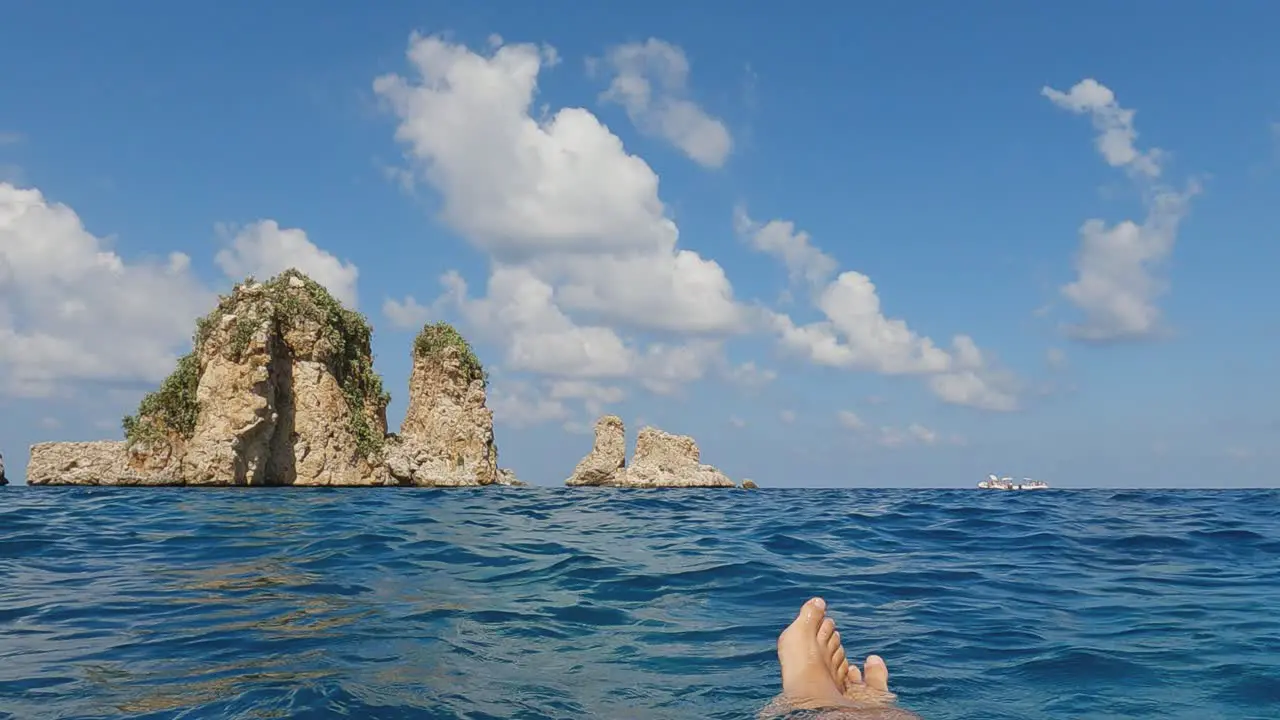 Personal low angle perspective of legs and feet floating on sea water with Scopello Stacks or Faraglioni in background