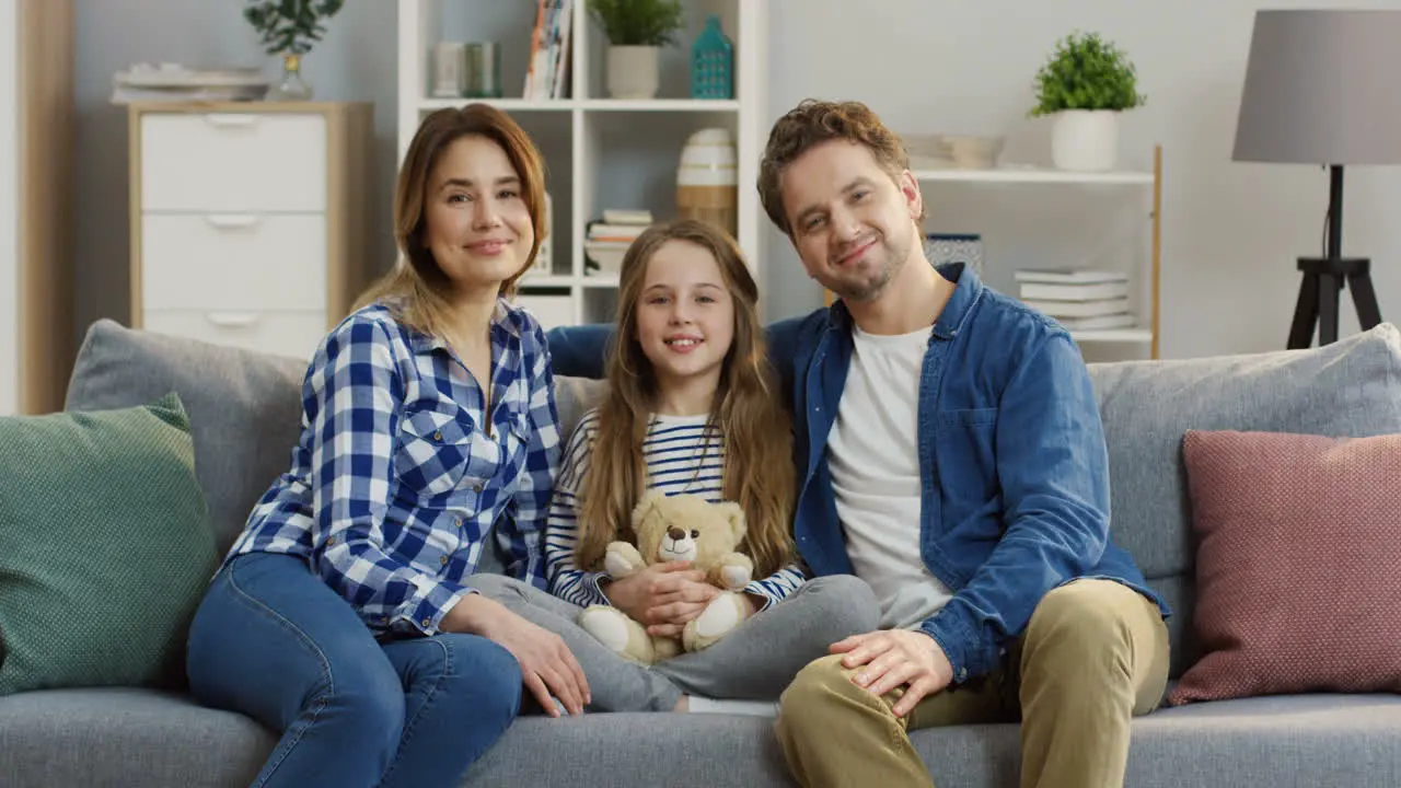 Portrait Shot Of The Young Happy Parents Sitting With Their Little Cute Daughter On The Couch In The Living Room Looking At Eacj Other And Smiling To The Camera