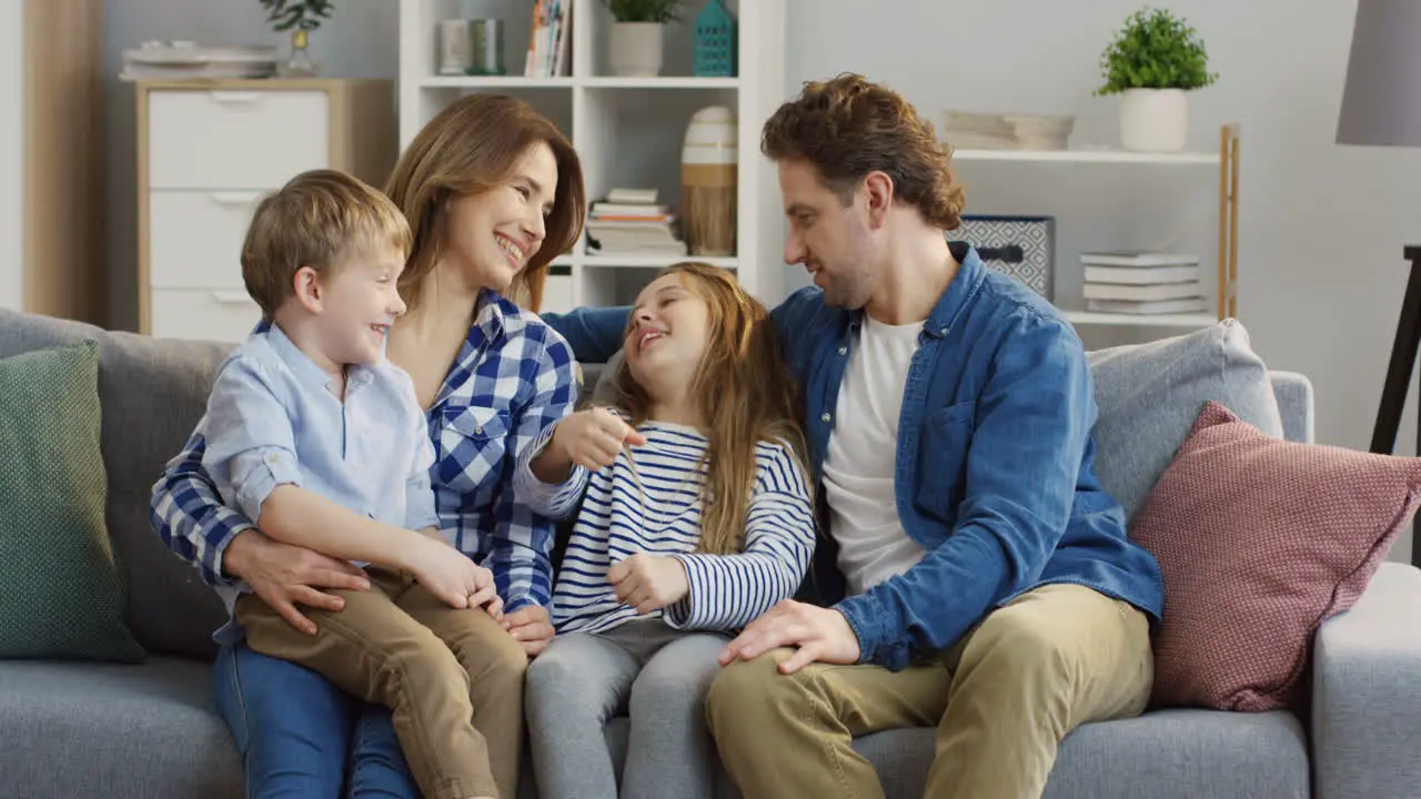 Happy Family Sitting On The Cozy Couch In The Living Room And Talking With Their Small Cute Kids Who Sitting On Their Knees And Between Them