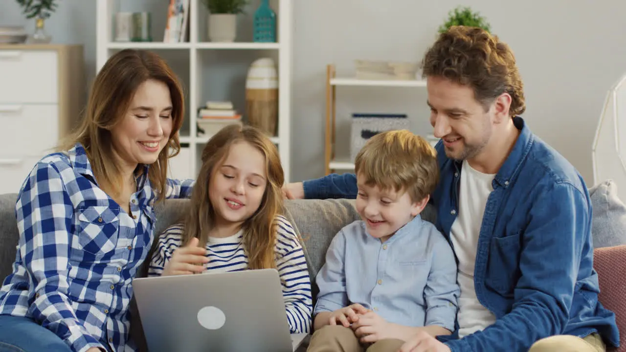 Joyful Young Parents With Little Daughter And Son Sitting On The Sofa In The Living Room And Talking While Having Videochat On The Laptop Computer