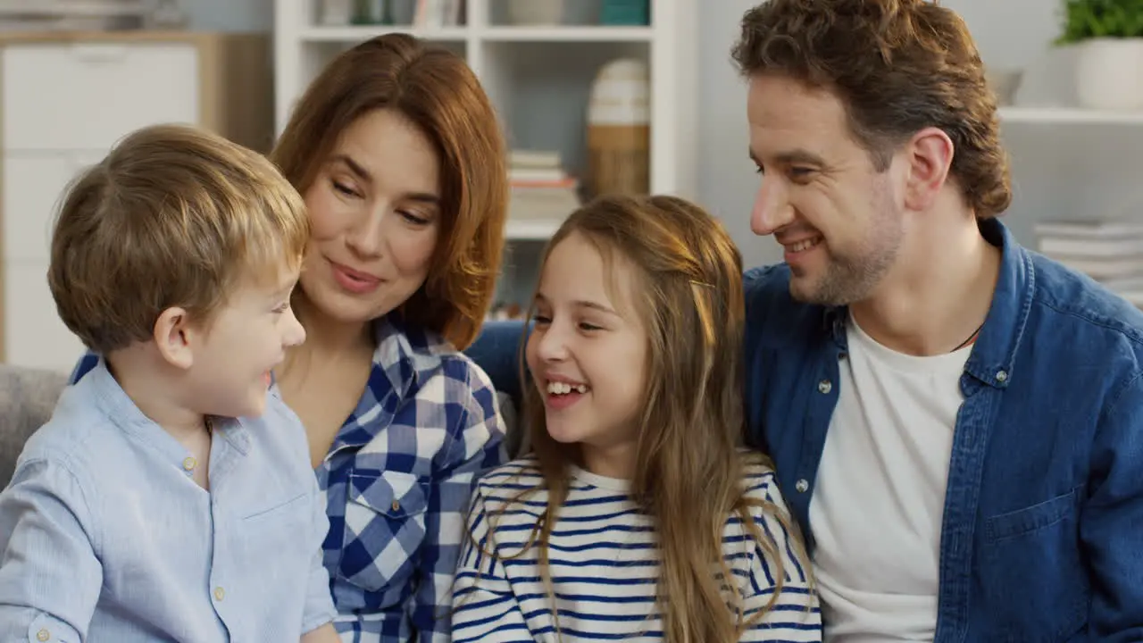 Close Up Of The Young Parents Sitting On The Sofa In The Living Room And Talking With Their Small Cute Daughter And Son Who Sitting On Their Knees