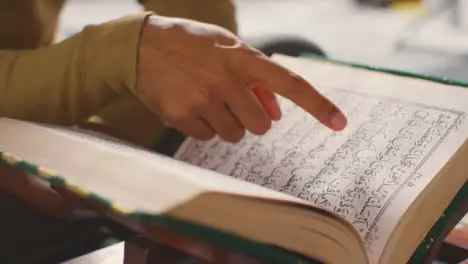 Close Up Of Open Copy Of The Quran On Stand At Home With Man Reciting Or Reading