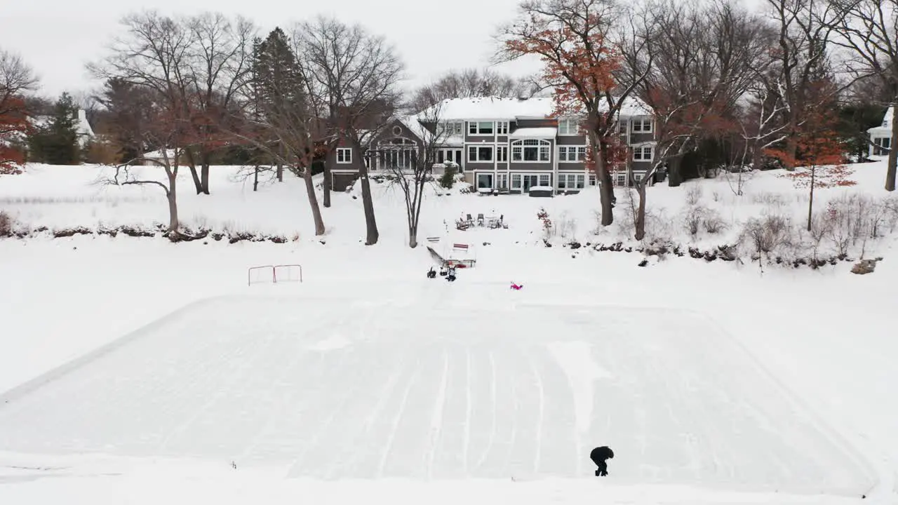 Aerial person clearing snow to make a small hockey ice rink on a frozen lake outside of house