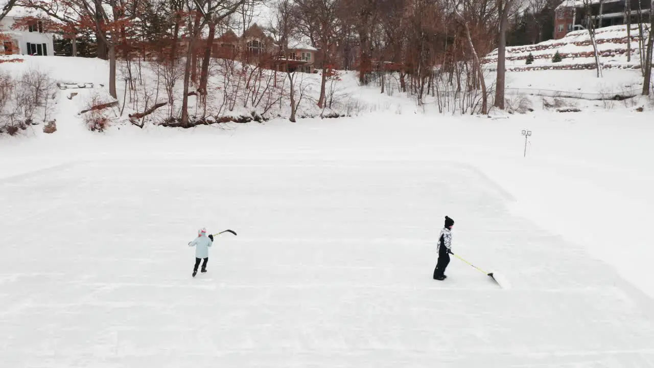 Aerial two kids clearing snow and ice skating on outdoor ice hockey rink on a frozen lake