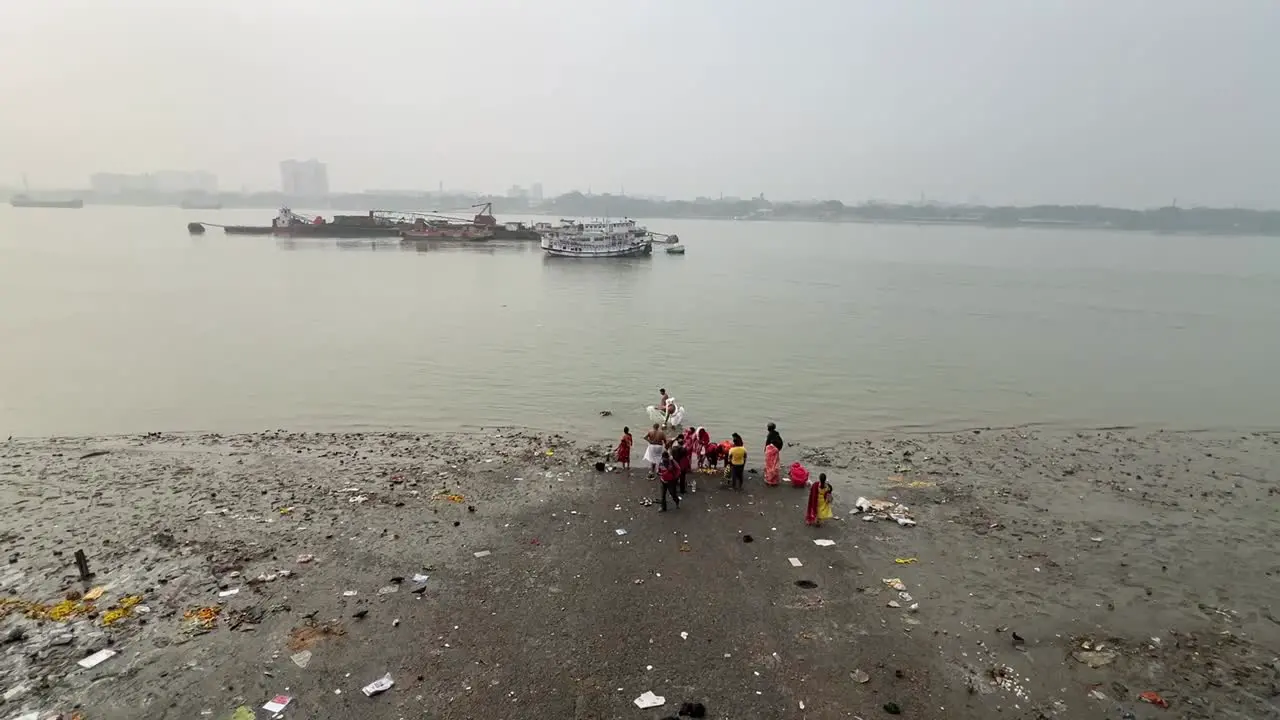 Rear view wide shot of people bathing and offering prayers to god near Hooghly river with cityscape at background in Kolkata India