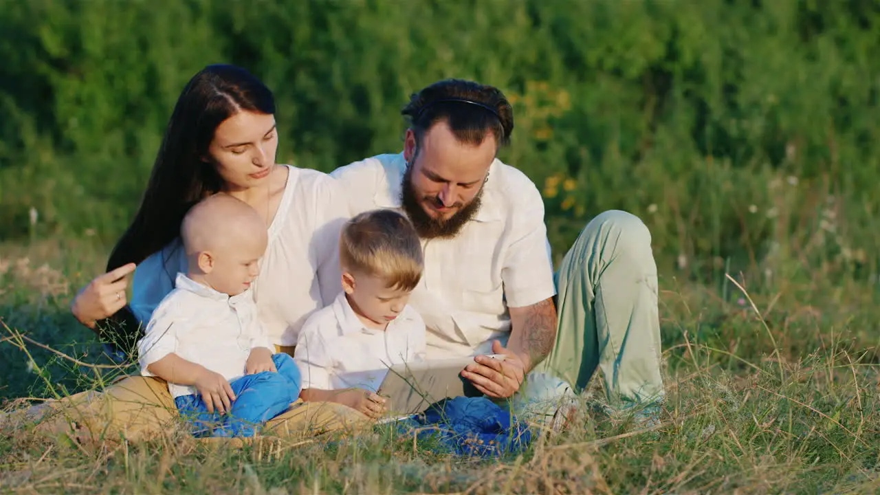 Two Small Brothers With Their Parents Play On A Tablet Among The Flowering Meadow