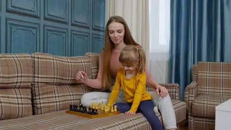 Small cute daughter child and young mother playing chess at home sofa leisure hobbies activities