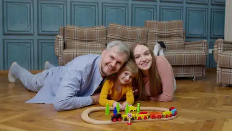 Young mother and father playing with child daughter riding toy train on wooden railroad game at home