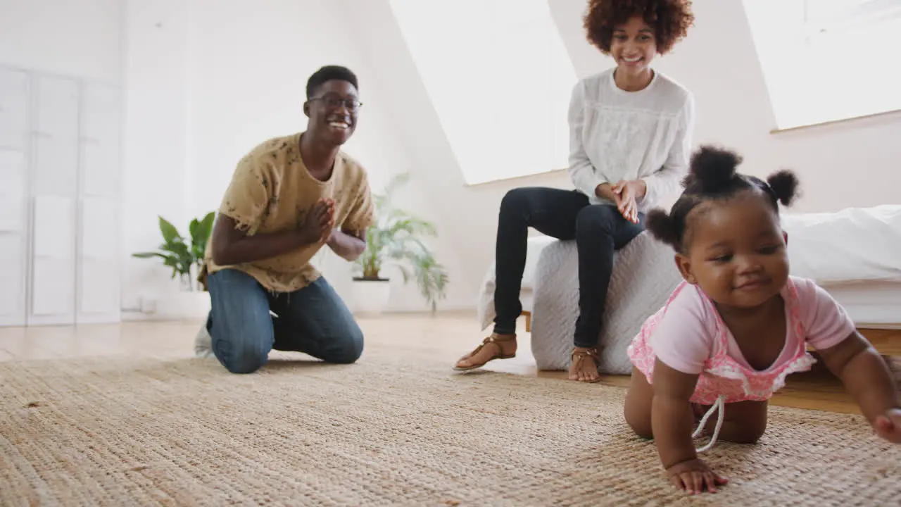 Baby Girl Learning To Crawl At Home As Parents Sit On Floor And Give Encouragement