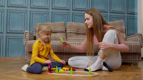 Mother playing with child kid daughter riding toy train on wooden railroad blocks board game at home