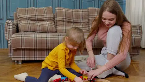 Mother with little daughter child girl riding toy train on wooden railway blocks board game at home