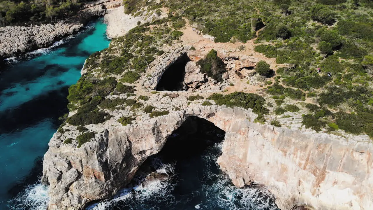 a drone shot flying backwards away from a sea arch with a couple of tourists entering the frame near cala varques in majorca