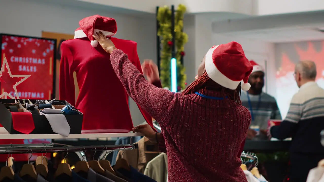 African american retail assistant wearing Santa hat helping asian client browsing through festive clothing in Christmas shopping store during winter holiday season Employee assisting customer