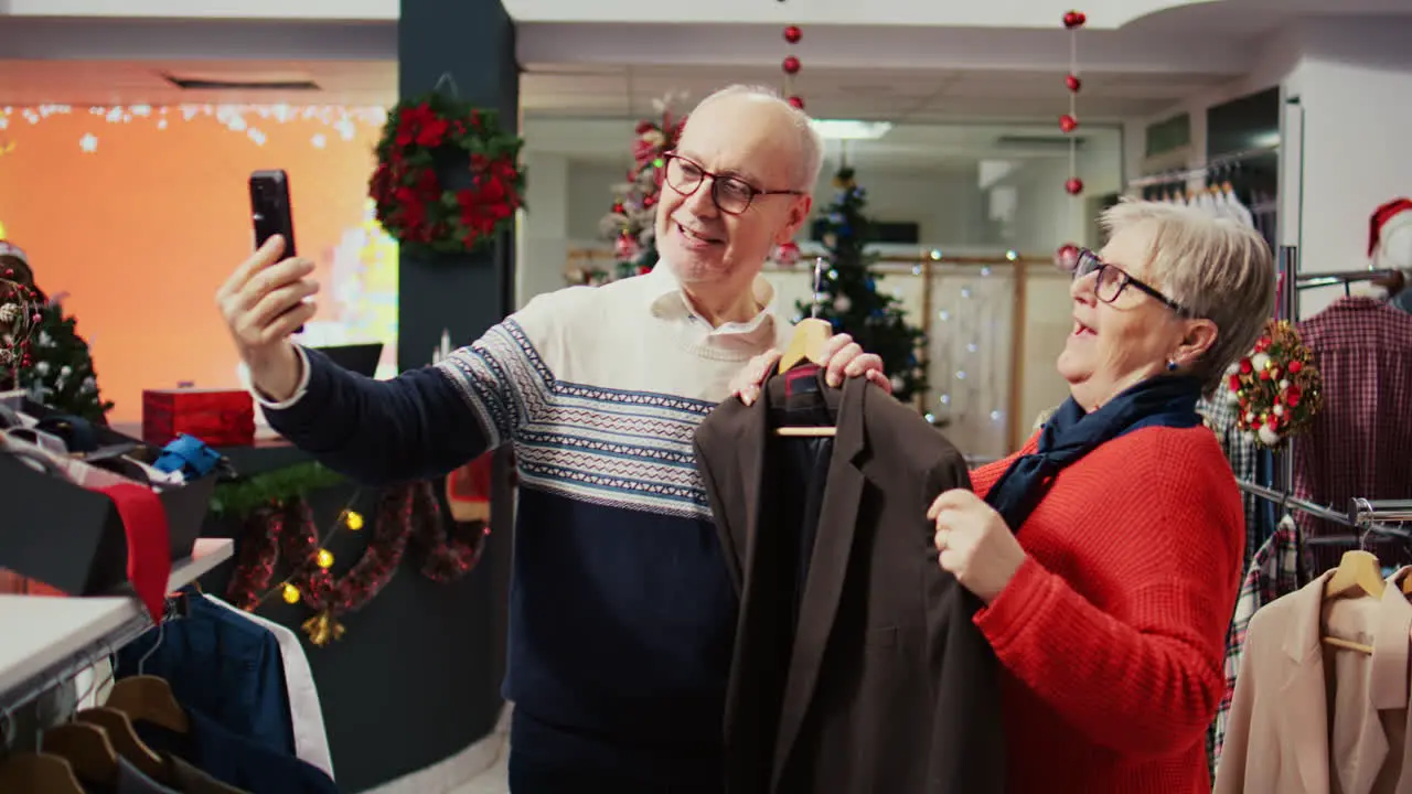 Elderly couple in xmas adorn shopping mall clothing store talking with grandson over internet videocall asking him to pick between stylish blazers to buy as present before Christmas family gathering