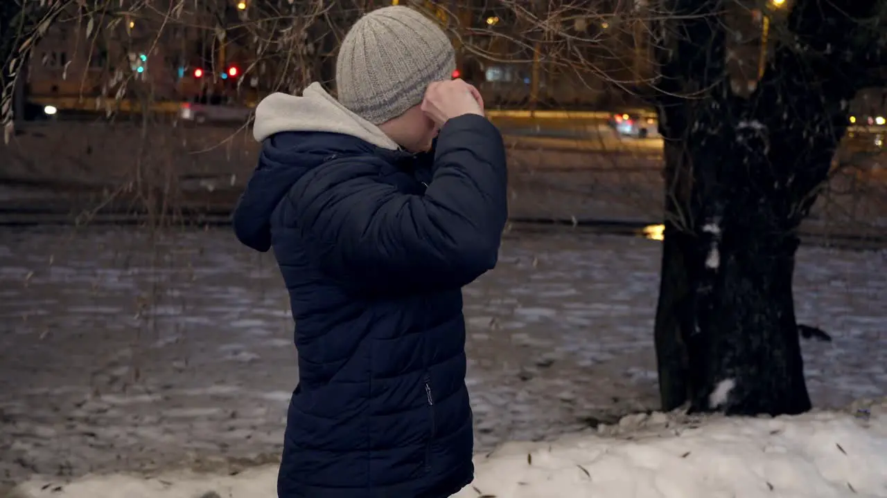 Caucasian man with blue jacket puts on a hat outdoors in cold winter evening