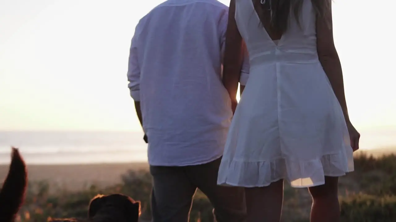 Slow motion handheld shot of a couple in love the woman dressed in a white dress her man dressed in white shirt with their dog during a romantic walk on the beach at a beautiful sunset