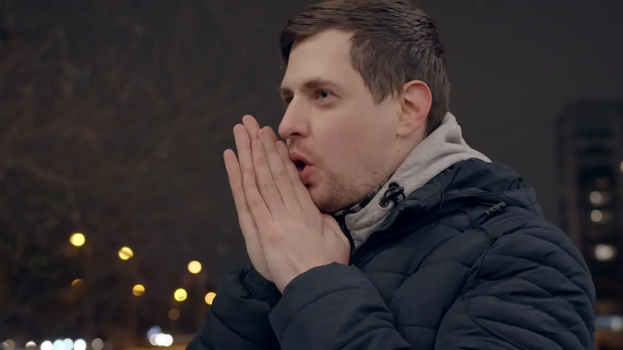 Frozen young man warming hands with breath cold night in winter handheld shot with copy space