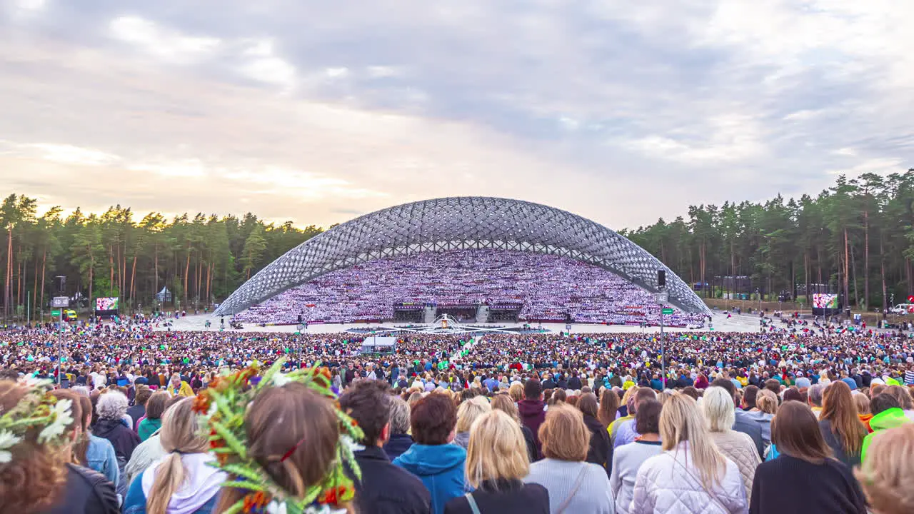 Timelapse shot of huge crowd of people watching Latvian Song and Dance Festival underway in an outdoor stadium on a cloudy day