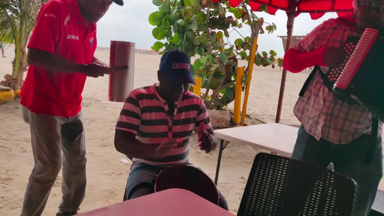 A man is playing the caja with his hands along his band members who play the Guacharaca and the accordion