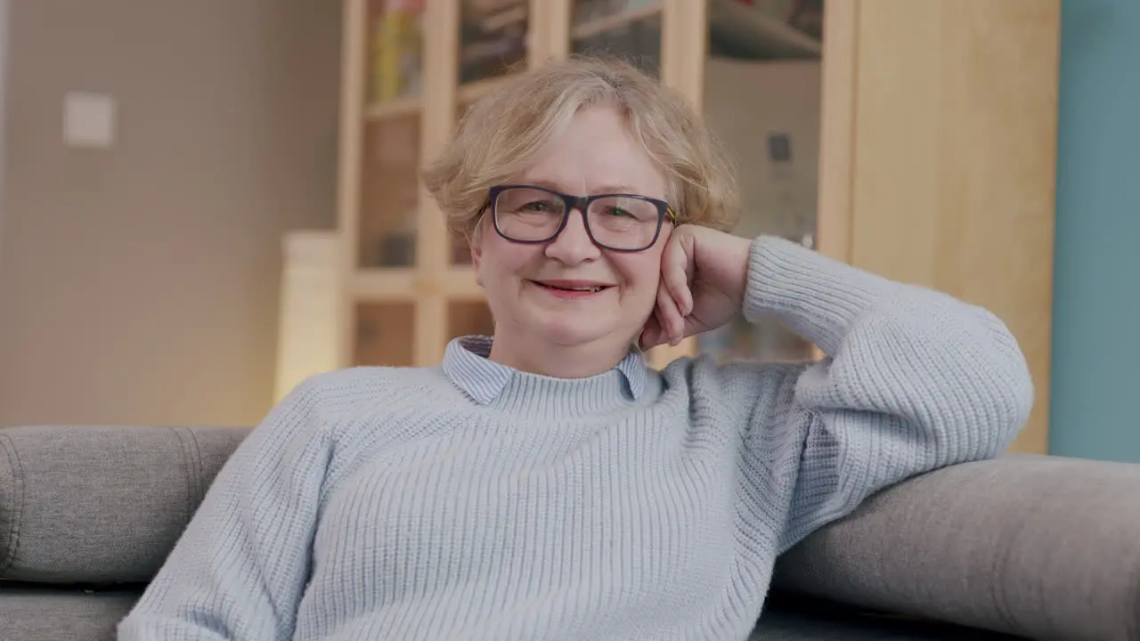 Smiling elderly caucasian woman with glasses sits relaxed on sofa frontal